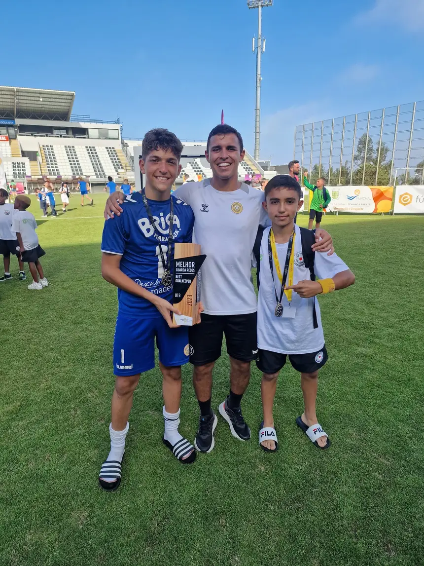 Treinador com dois jovens jogadores no relvado, um deles com um troféu nas mãos e uma medalha ao pescoço, com as bancadas ao fundo.