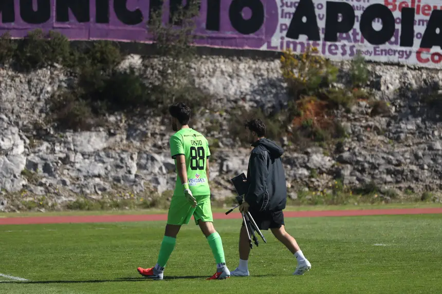 Guarda-redes a sair do relvado acompanhado por um elemento da equipa técnica, que leva o tripé dobrado.