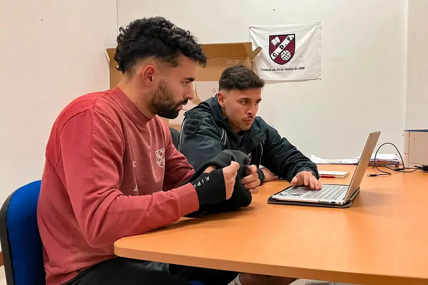 Dois elementos da equipa técnica a analisar vídeo num portátil, numa sala do clube com a bandeira na parede.
