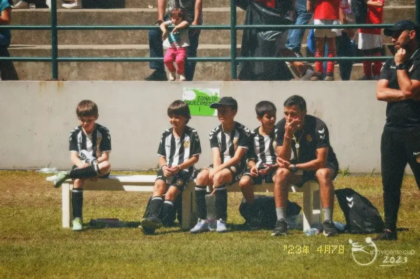 Jovens jogadores sentados no banco à beira do campo com o treinador, num torneio.